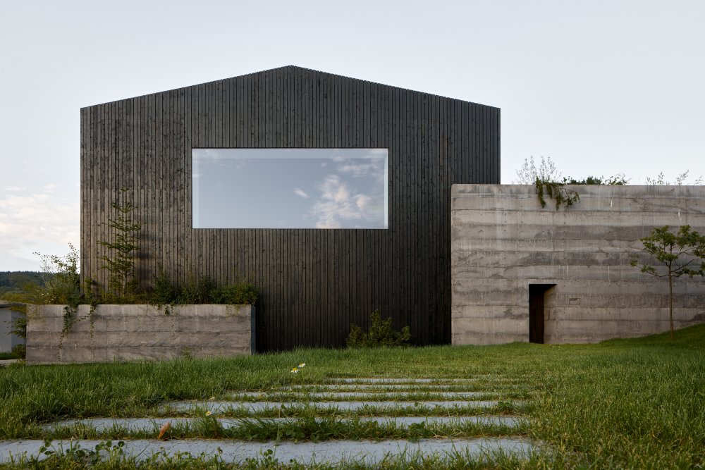 Vnitřní prostor formuje otevřený krov, zapuštěné balkony a středová chodba, která propojuje celý dům. Po jejích stranách se rozprostírají kanceláře a zasedací místnosti, zatímco centrální část funguje jako společné náměstí – místo setkávání a sdílení. Skrze posuvnou skleněnou stěnu se otevírá do zahrady a dál do louky.