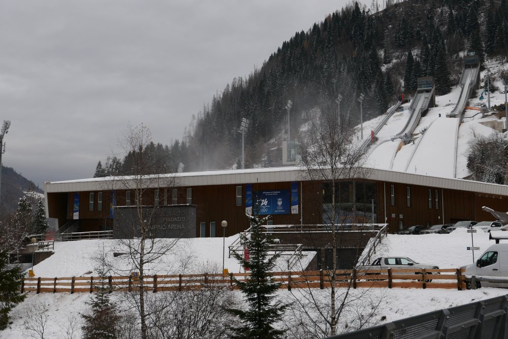 Rekonstrukce skokanského areálu v Predazzu pro Zimní olympijské hry Milano-Cortina 2026 představuje citlivý kompromis mezi sportovním prestiží, tlakem na udržitelnost a obrovskými veřejnými investicemi. Z ekonomického pohledu se projekt pohybuje v desítkách milionů eur