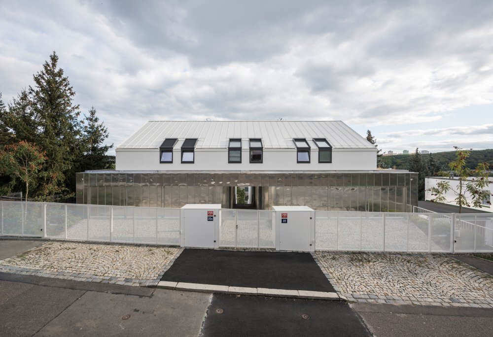 Výsledný dvojdům charakterizují terasy orientované na jih umožňující po svém obvodu výsadbu vyšší zeleně. Hmota je řešena jako ustupující bílý blok kopírující terén. Na bočních fasádách jsou dle výhledů z interiéru komponovány různé typy čtvercových oken. Na úrovni ulice je tento bílý monolit protnut horizontálním objemem garáží opláštěným přírodním hliníkovým falcovaným plechem.