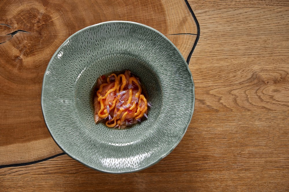 La Degustation Bohême Bourgeoise nabízí moderní fine dining inspirovaný českou a středoevropskou kuchyní. Na snímku je pstruh pošírovaný v šunkovém sádle s bobkovým listem a kořením (71 °C, 7–8 min). Omáčka z čerstvé rajské šťávy, tymiánu, šalotky a másla; majonéza z pečených rajčat, olej ze sušených rajčat a pažitkové květy z Kytína.