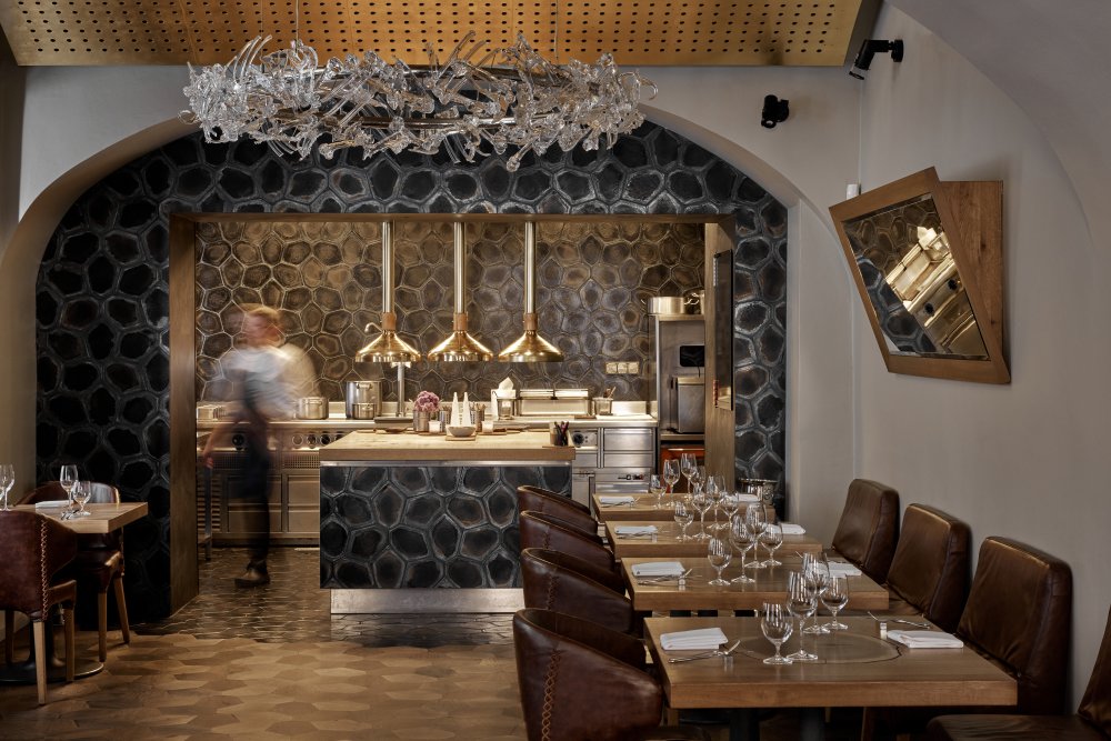 La Degustation Bohême Bourgeoise ukazuje, jak může špičková gastronomie pracovat s českou tradicí současným jazykem. Interiér studia Olgoj Chorchoj propojuje křišťál, dub, čedič i kůži a vytváří prostor, který nepůsobí okázale, ale soustředěně a sebevědomě. Dominantou je autorský lustr Consommé, jehož skleněné prvky odkazují k práci se surovinou i respektu k řemeslu.