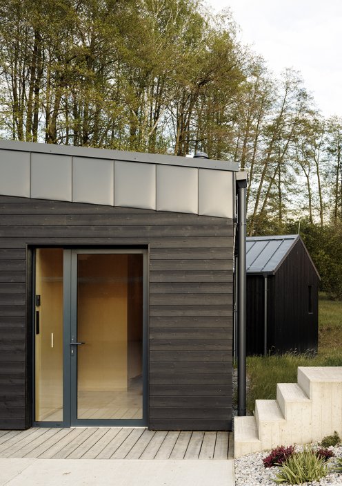 Černá dřevěná fasáda kontrastuje se světlou zelení pozemku a podtrhuje jednoduchou geometrii celé stavby.