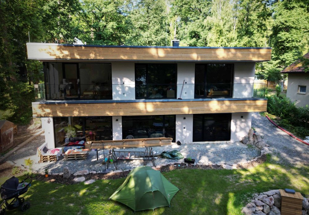 V jevanském lese vznikl rodinný dům, který se z ulice téměř ztrácí, zatímco směrem do zahrady se otevírá velkými prosklenými plochami do zeleně. Architekti Tomáš Hladík a Antonín Hůla pracovali se svažitým terénem, jednoduchými bílými plochami a dřevem, které dům propojuje s okolní krajinou. Část nábytku navíc vznikla ze stromů, jež původně rostly přímo na pozemku.