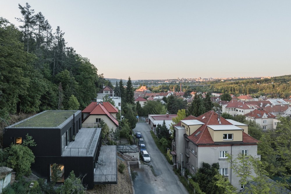 Na strmém pozemku pod lesem na Zbraslavi vznikl dům pro dvě spřátelené rodiny. Parcela s původní dřevěnou chatou byla obtížně zastavitelná, její cena ale podle autorů odpovídala zhruba osmdesátimetrovému bytu v lokalitě. Architekti z kanceláře Mimosa na ni posadili dva jednoduché objemy, každý s vlastním bytem, terasou a výhledem do Břežanského údolí. Dům díky tmavé fasádě splývá s lesem a využívá společné technické zázemí, tepelné čerpadlo, dešťovou vodu i vegetační střechu.